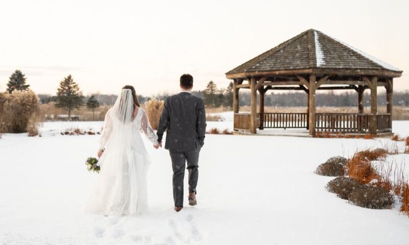 Bride and groom walking hand in hand through the snow toward a wooden gazebo on a winter day.