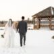 Bride and groom walking hand in hand through the snow toward a wooden gazebo on a winter day.