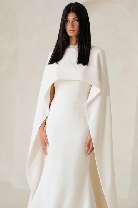 Woman with long straight black hair wearing a sleek, white gown with a structured cape overlay, standing against a neutral background.