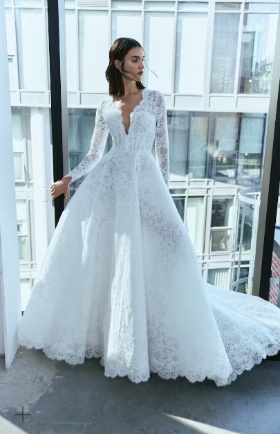 Bride wearing a long-sleeved lace wedding gown with a deep V-neckline and full skirt, standing by a large window with modern buildings outside.