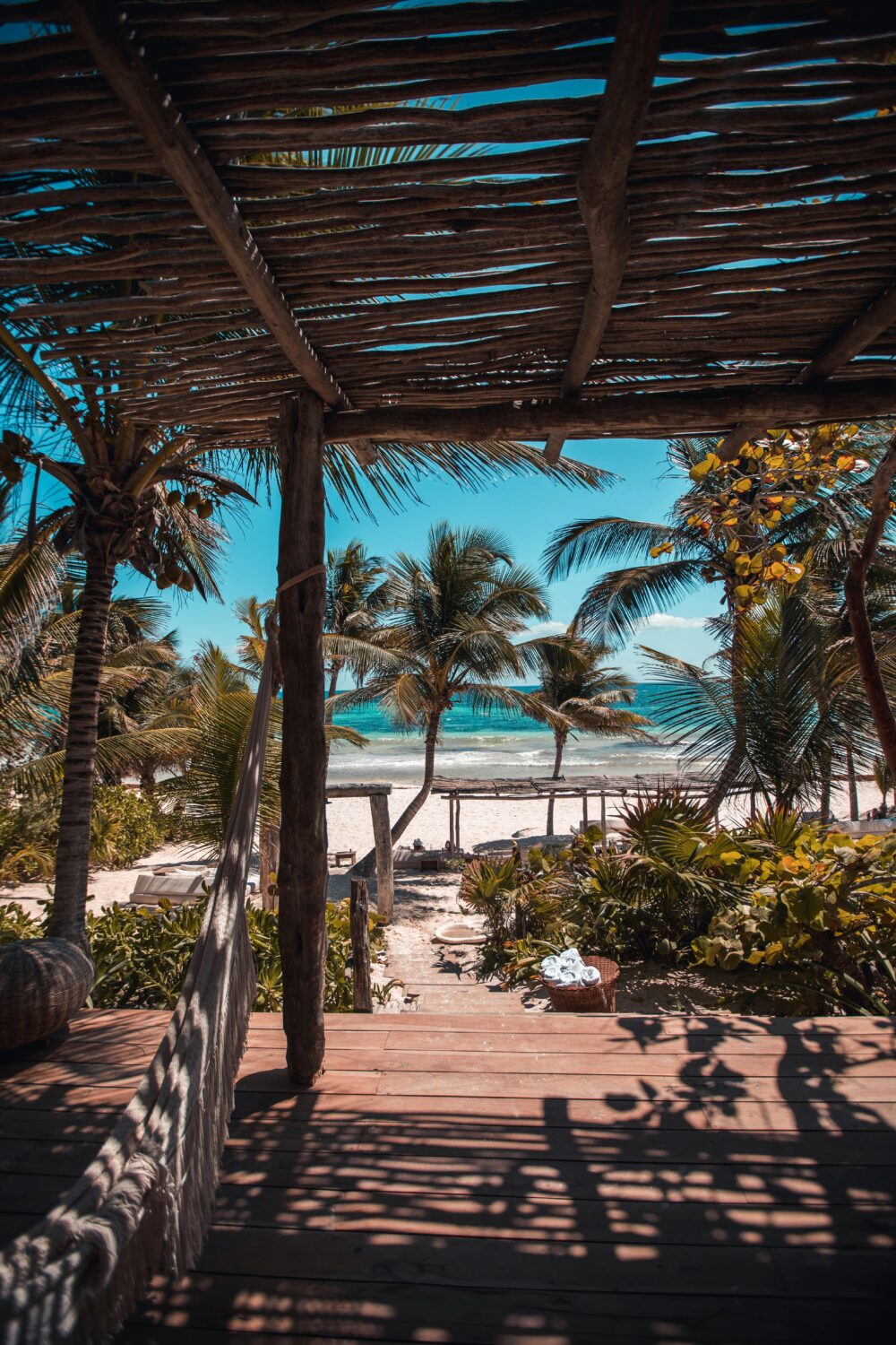 View of a tropical beach with palm trees, clear turquoise water, and white sand seen from under a wooden pergola.