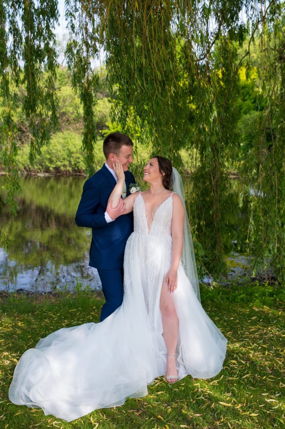 A bride and groom standing under the shade of willow tree branches beside a pond, looking at each other lovingly.