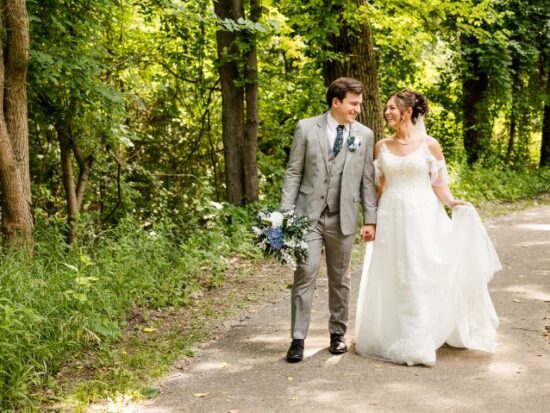 A bride and groom walking along a wooded path, smiling warmly at each other while holding hands, with the groom carrying a bouquet of blue and white flowers.