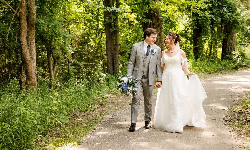 A bride and groom walking along a wooded path, smiling warmly at each other while holding hands, with the groom carrying a bouquet of blue and white flowers.