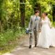 A bride and groom walking along a wooded path, smiling warmly at each other while holding hands, with the groom carrying a bouquet of blue and white flowers.