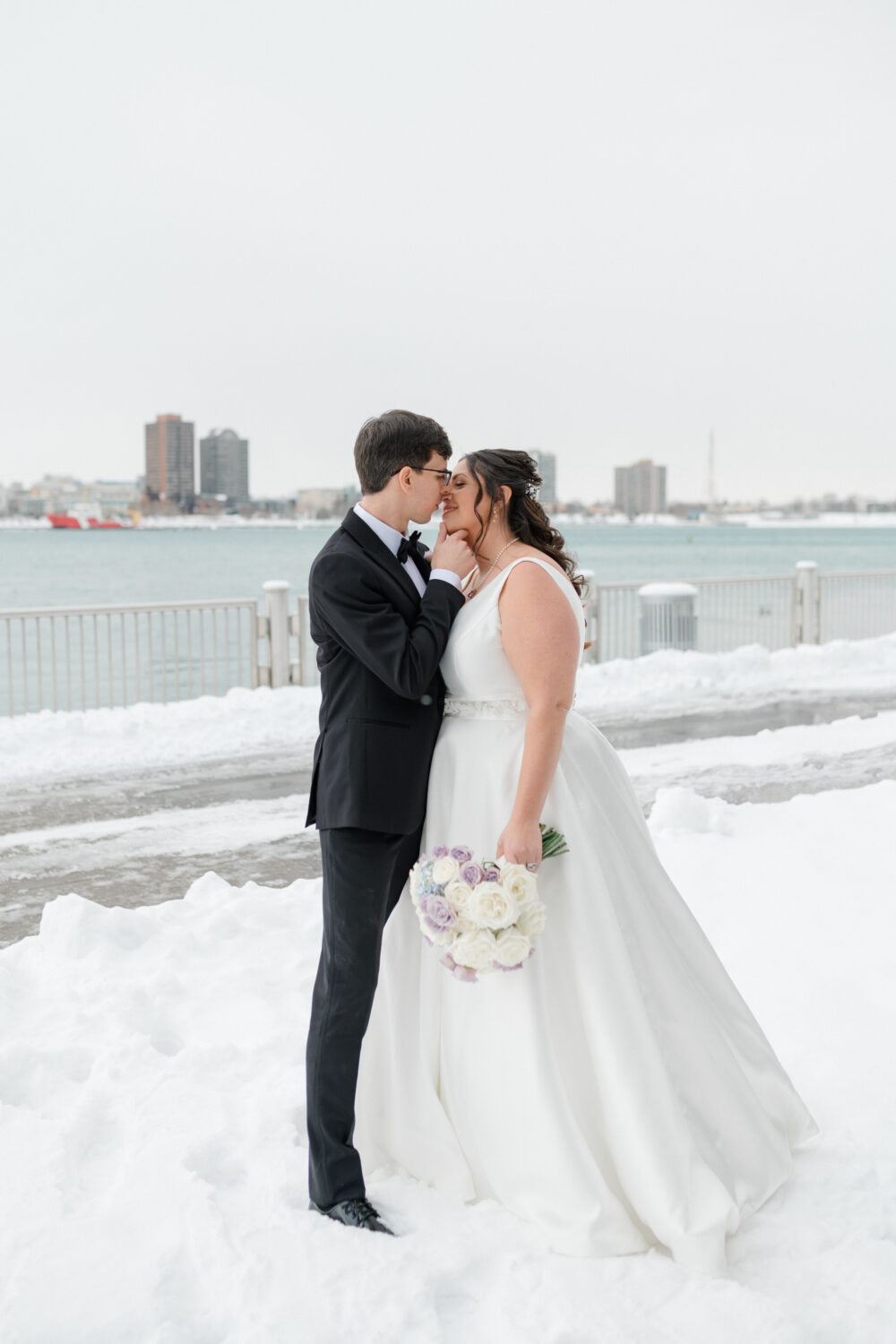 Groom gently holding bride’s chin as they share a romantic moment in the snow by a waterfront.