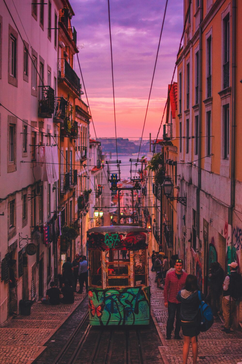 Narrow street in a city with a graffiti-covered tram, people walking, and a purple-pink sunset over water in the background.