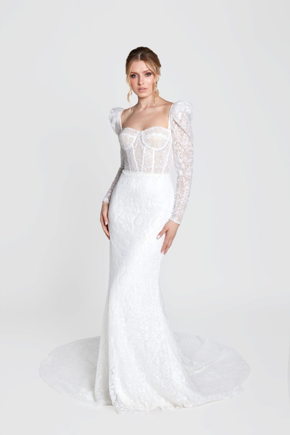 Bride in a fitted lace wedding dress with long puffed sleeves and a sweetheart neckline, looking forward against a plain light background.
