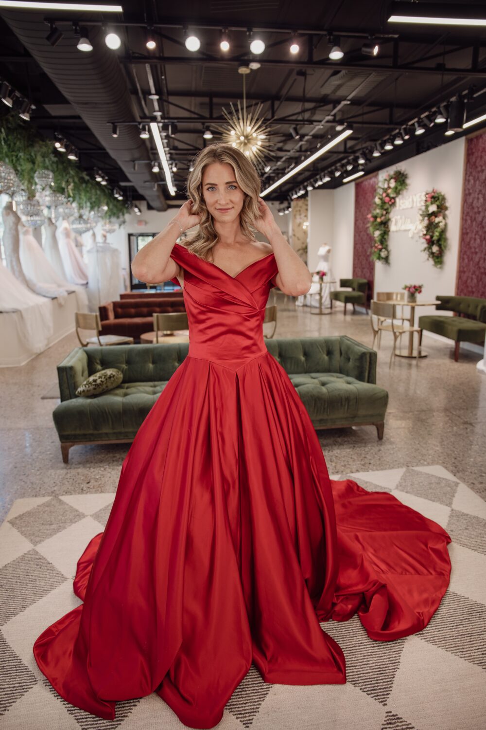 Woman wearing an off-the-shoulder red satin gown standing in a bridal boutique.