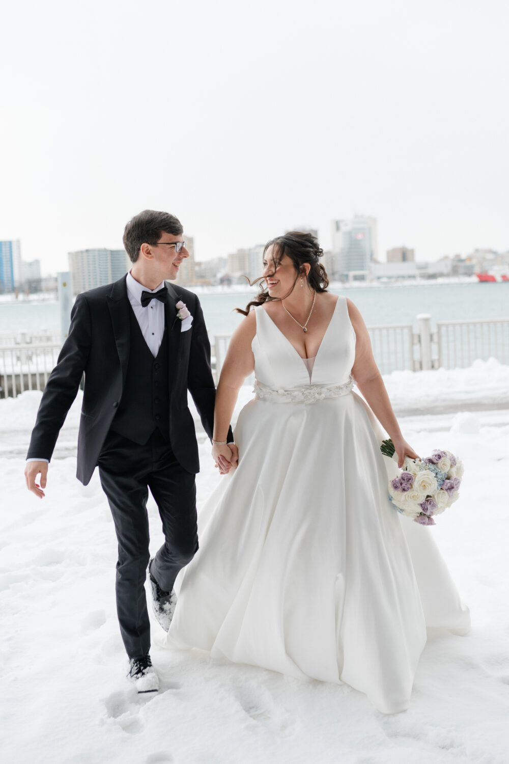 Bride and groom smiling at each other while walking hand in hand through the snow by a waterfront.