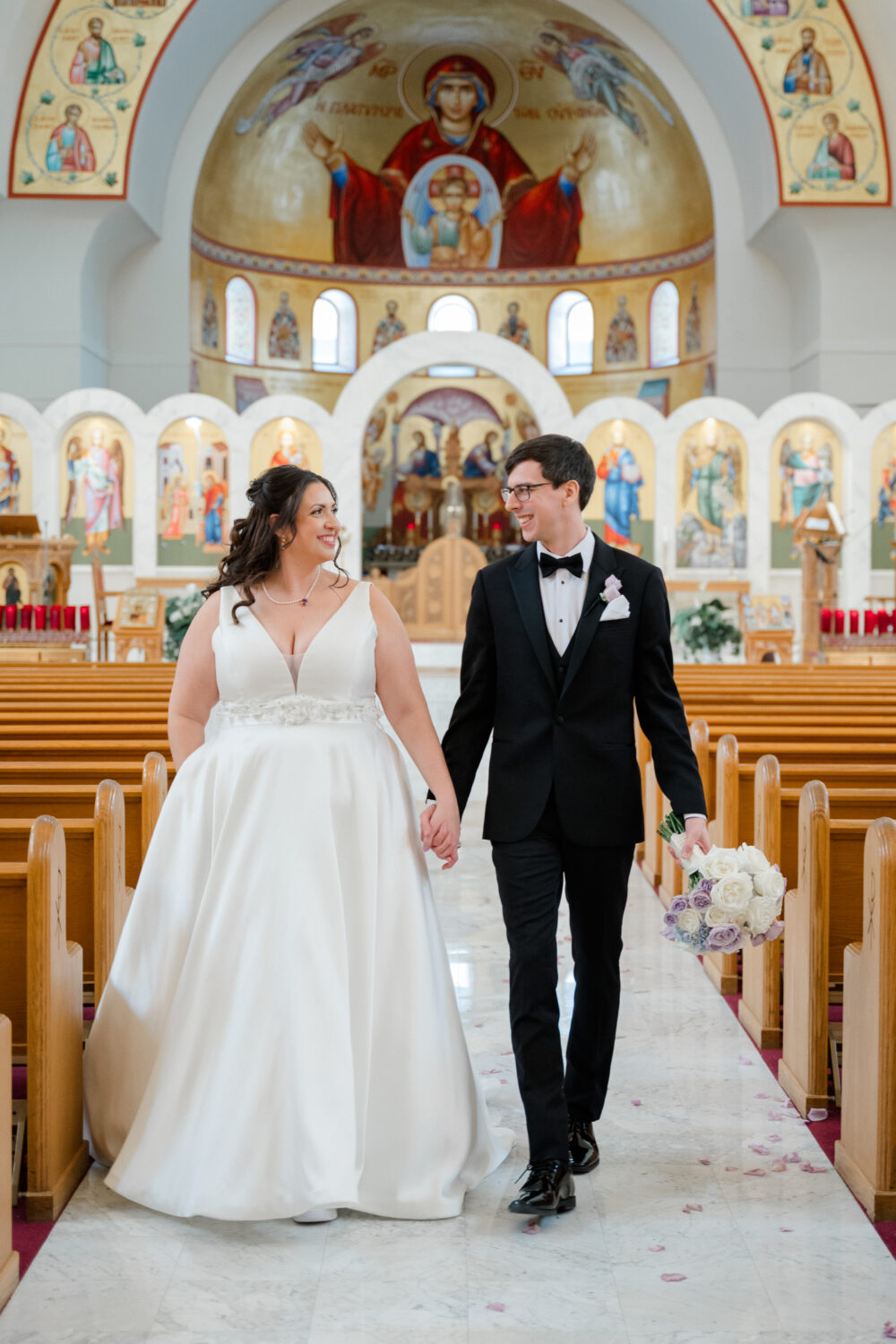 Bride and groom holding hands while walking down the aisle of a church adorned with religious artwork.