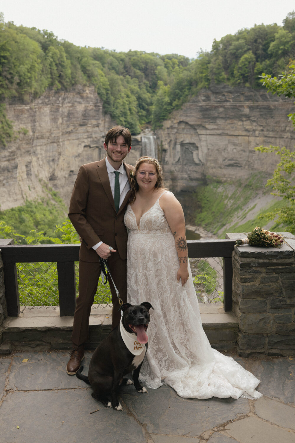 A smiling bride and groom standing together in front of a scenic waterfall, with their black and white dog wearing a bandana sitting at their feet.