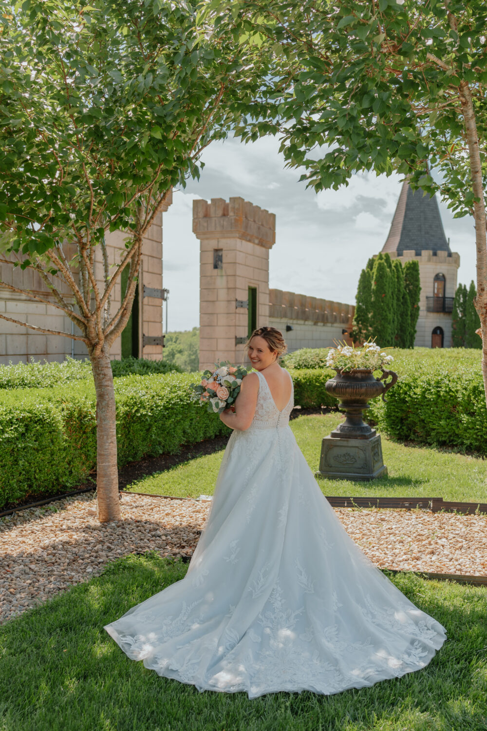 A bride in a long white gown holding a bouquet and looking over her shoulder while standing in a garden with castle-like stone walls and towers behind her.
