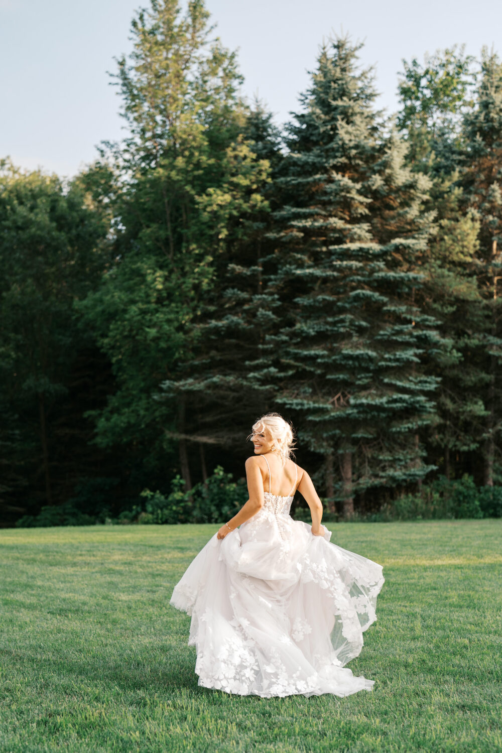 A bride in a white lace wedding dress running joyfully across a grassy field, holding her skirt up, with tall pine trees in the background.
