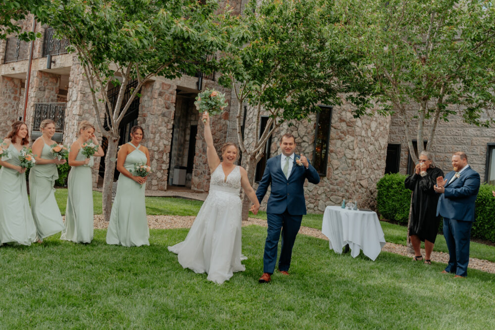 A bride and groom walking hand in hand outdoors, the bride raising her bouquet in celebration, surrounded by bridesmaids in sage green dresses and groomsmen applauding.