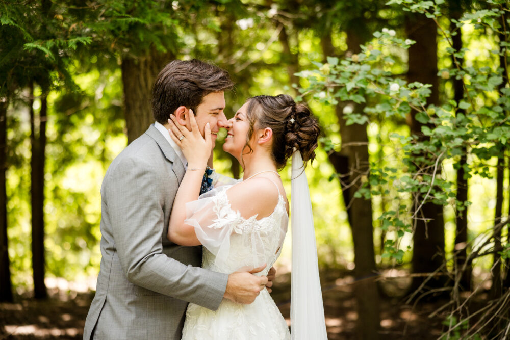 A bride and groom standing close in a wooded area, smiling and touching noses as they hold each other affectionately.