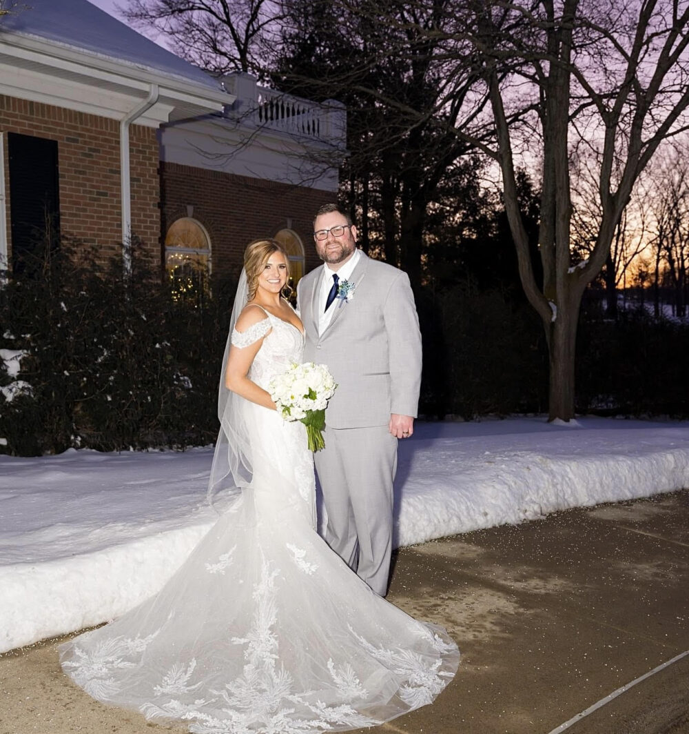 Bride in an off-the-shoulder lace gown standing beside groom in a light gray suit on a snowy evening.