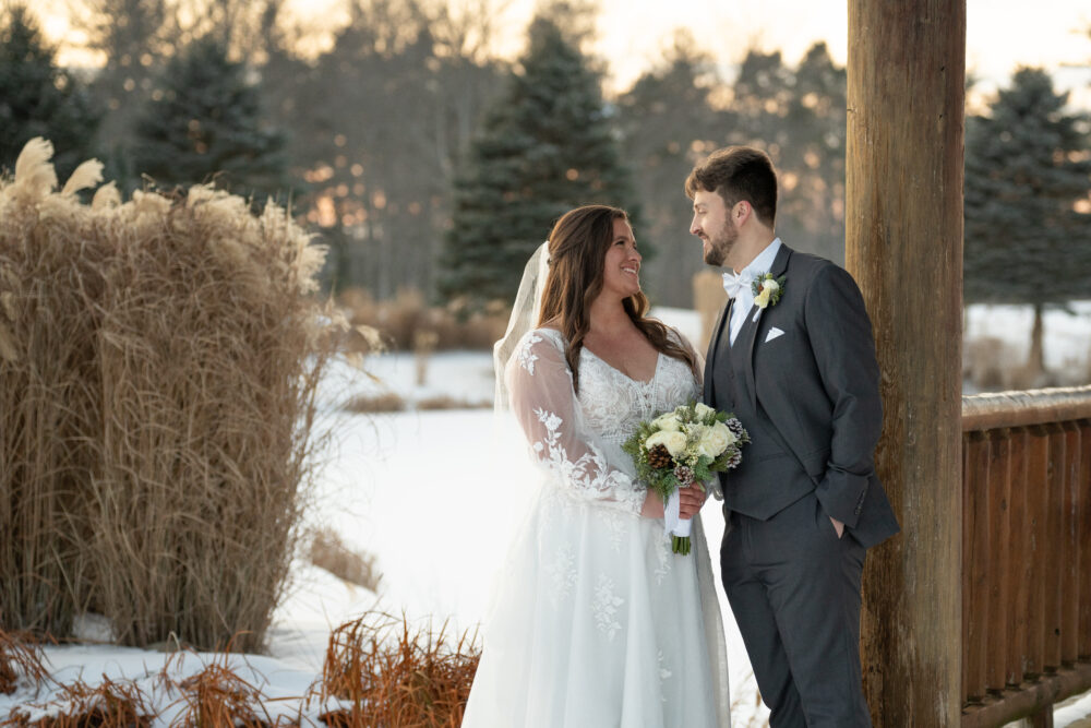 Bride in a long-sleeve lace gown holding a bouquet and smiling at groom by a wooden gazebo in winter.