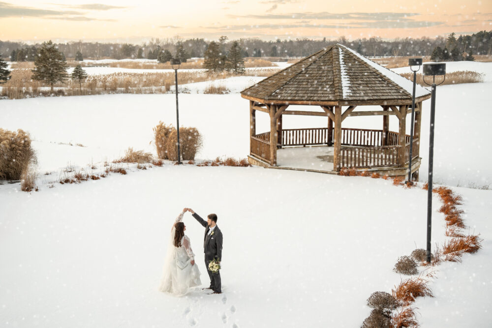 Bride and groom dancing together in the snow near a wooden gazebo at sunset.