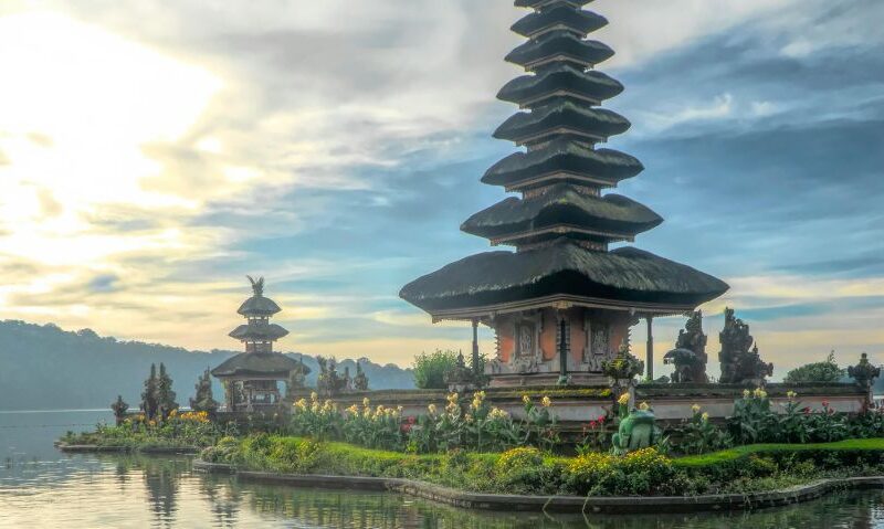 A temple on an island off the shore with the sun rising on the horizon in the background.