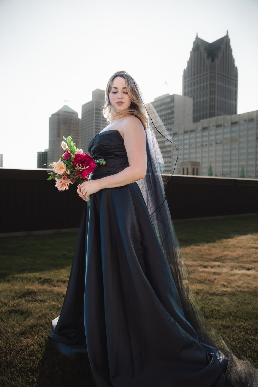 A woman in a strapless black gown and black veil holds a bouquet of bright flowers while standing on a rooftop with Detroit skyscrapers in the background.