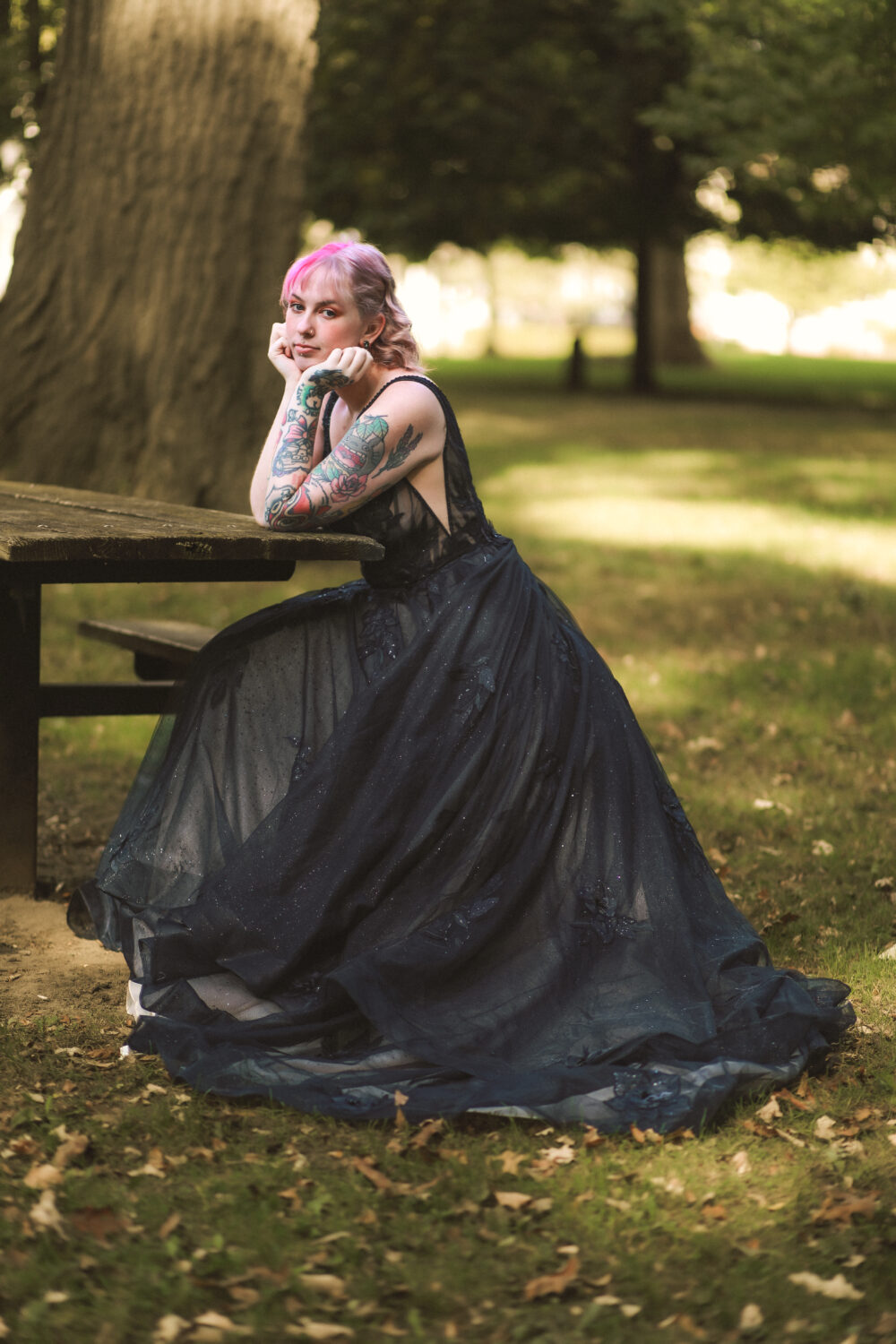 A woman with pink hair and tattooed arms wears a flowing black gown while sitting at a wooden picnic table in a park.