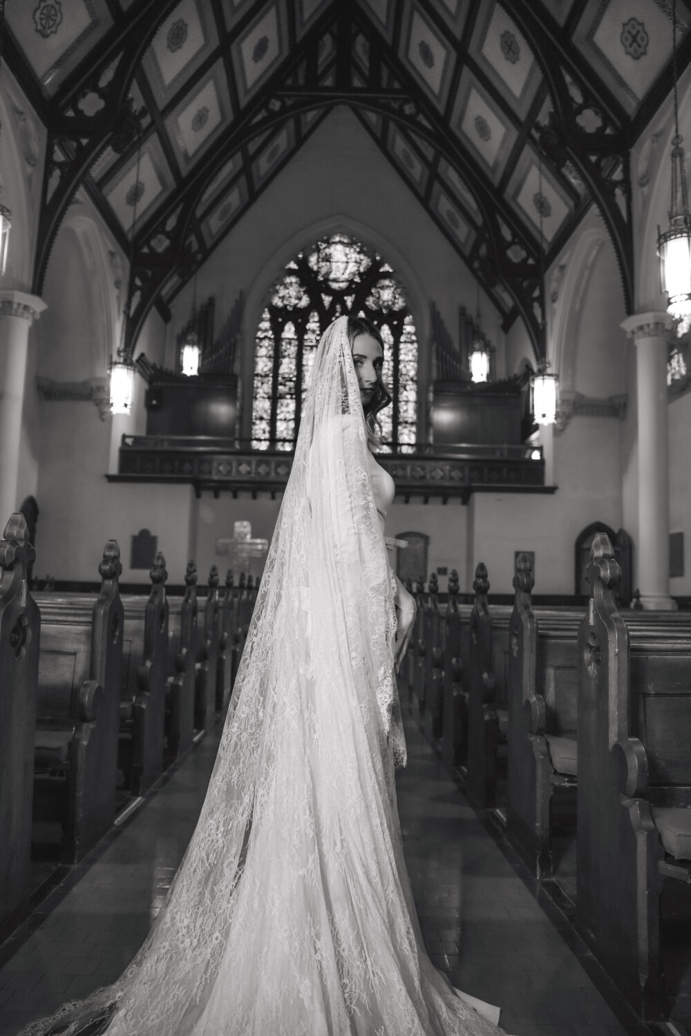 A bride in a long lace gown and cathedral veil stands in the aisle of a church, with stained glass windows visible behind her.