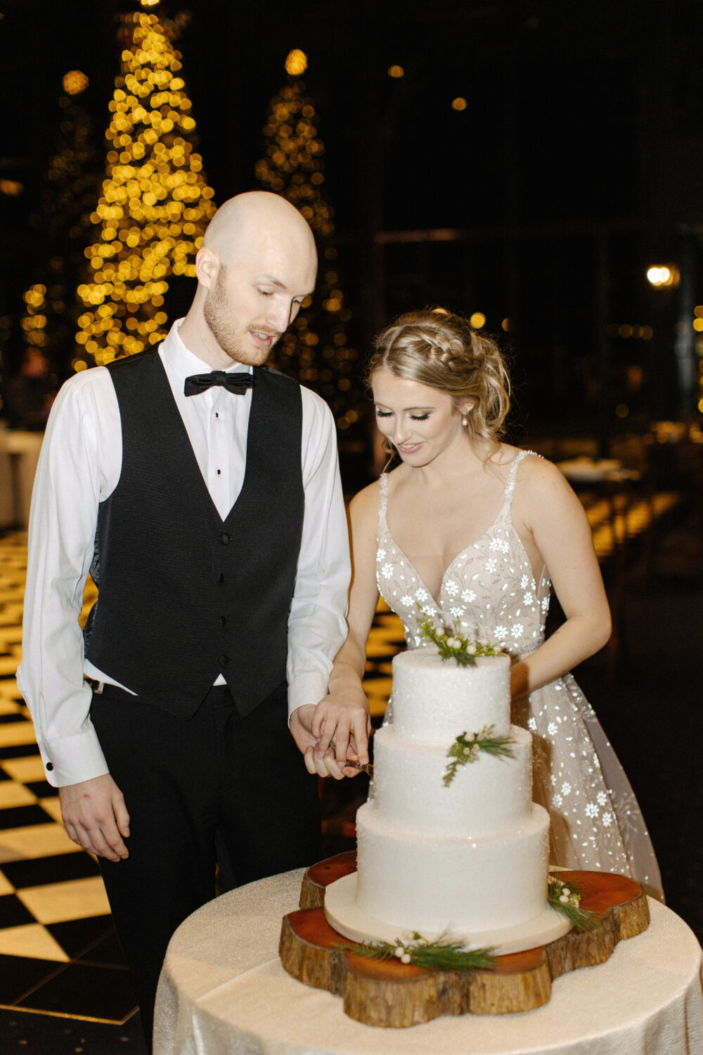 A bride and groom cut a three-tier white wedding cake decorated with greenery, with glowing Christmas trees in the background.