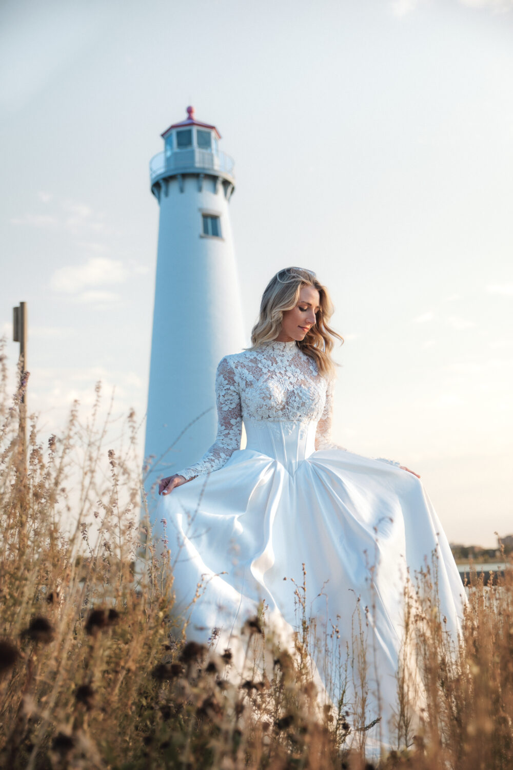 A bride in a lace long-sleeve gown poses outdoors with a lighthouse in the distance.