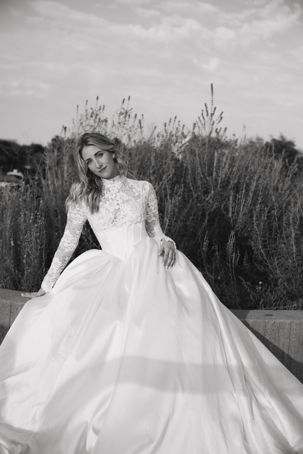 A bride in a lace long-sleeve gown with a full satin skirt sits gracefully near tall grasses outdoors.
