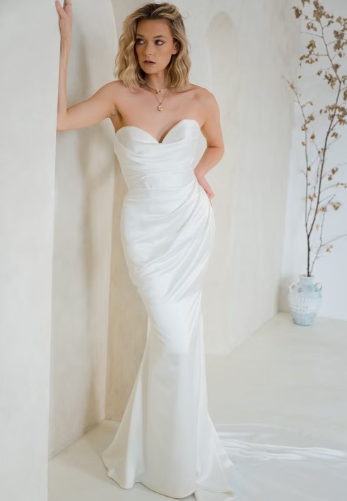 A bride in a strapless satin gown with a sweetheart neckline leans against a wall in a softly lit room.