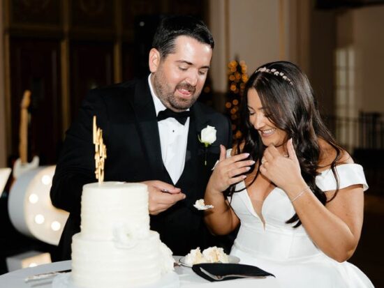 A bride and groom laugh while feeding each other wedding cake beside a two-tier white cake with a gold topper.