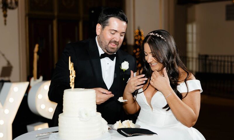 A bride and groom laugh while feeding each other wedding cake beside a two-tier white cake with a gold topper.