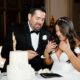 A bride and groom laugh while feeding each other wedding cake beside a two-tier white cake with a gold topper.