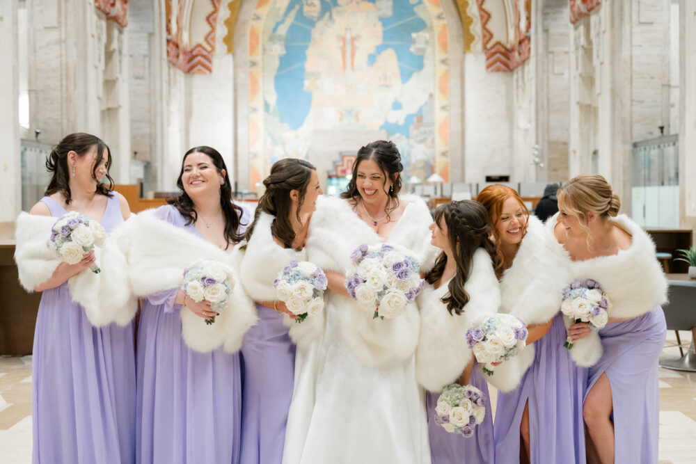 A bride in a white gown stands with her bridesmaids in lilac dresses, all holding bouquets and draped in white fur shawls.