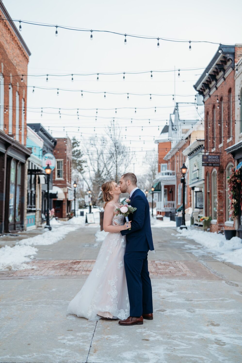 A bride and groom kiss on a snow-covered street lined with brick buildings and shops, beneath string lights.