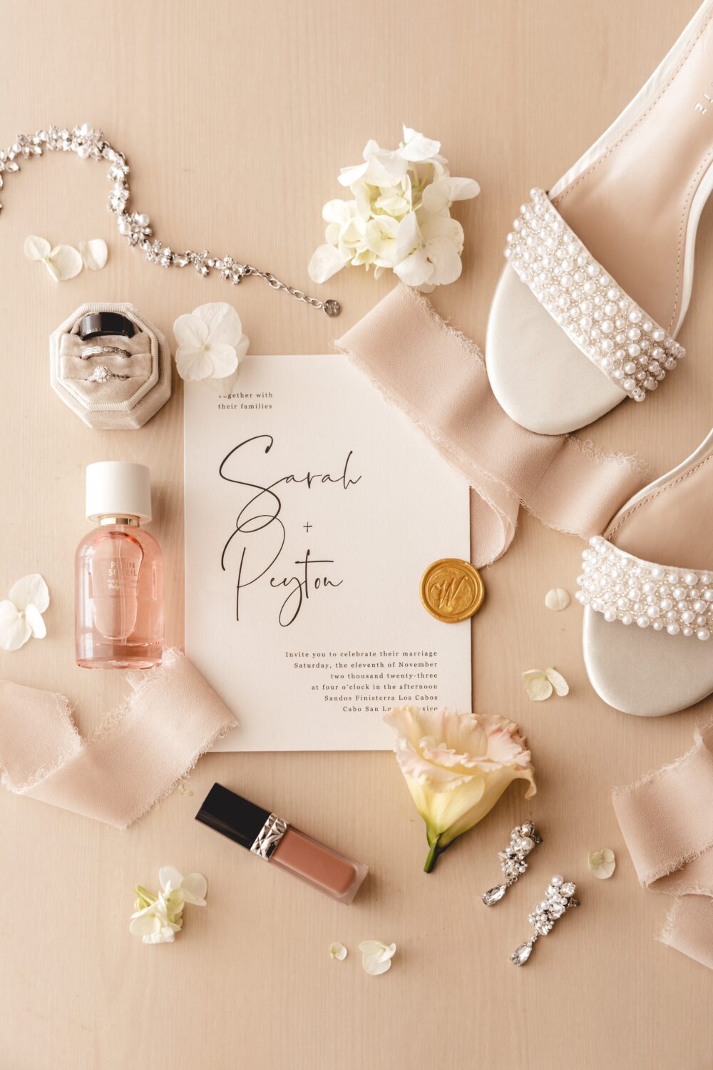 A flat lay of a wedding invitation surrounded by pearl bridal shoes, a perfume bottle, lipstick, earrings, a bracelet, and a small flower.