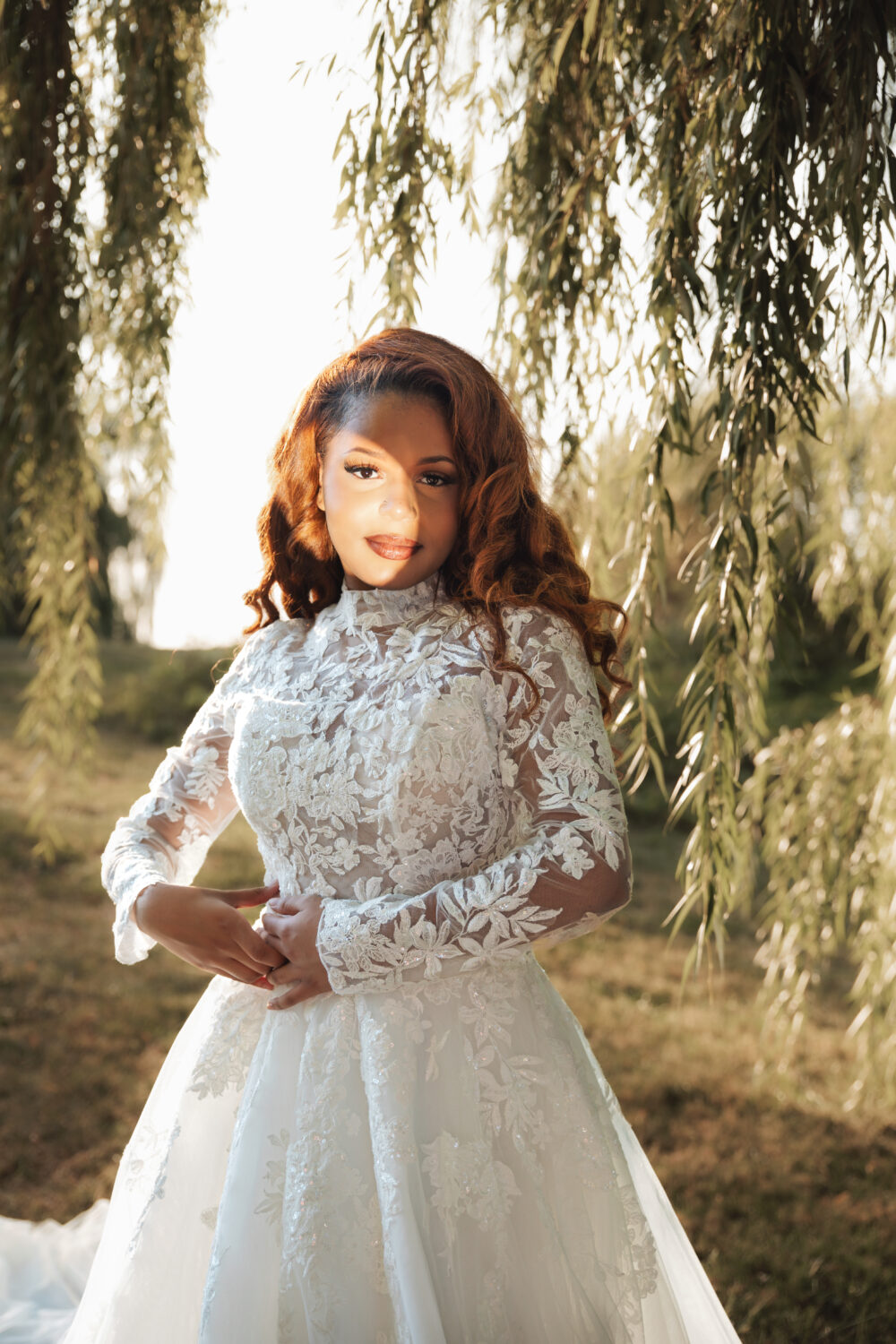 A bride in a lace long-sleeve gown stands under willow branches with sunlight highlighting her face.