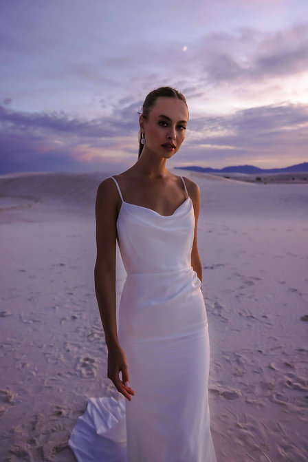 A bride in a sleek satin gown with thin straps poses against a desert landscape at sunset.