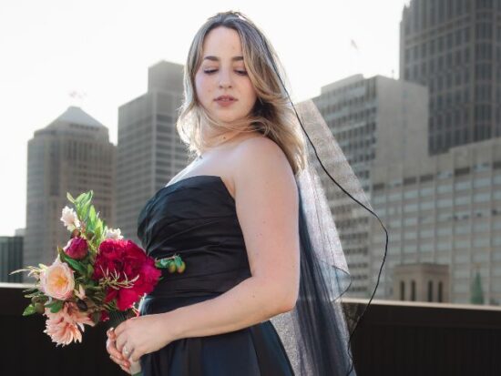 A woman in a black strapless gown and veil holds a bouquet of pink and red flowers while standing on a rooftop with tall buildings in the background.
