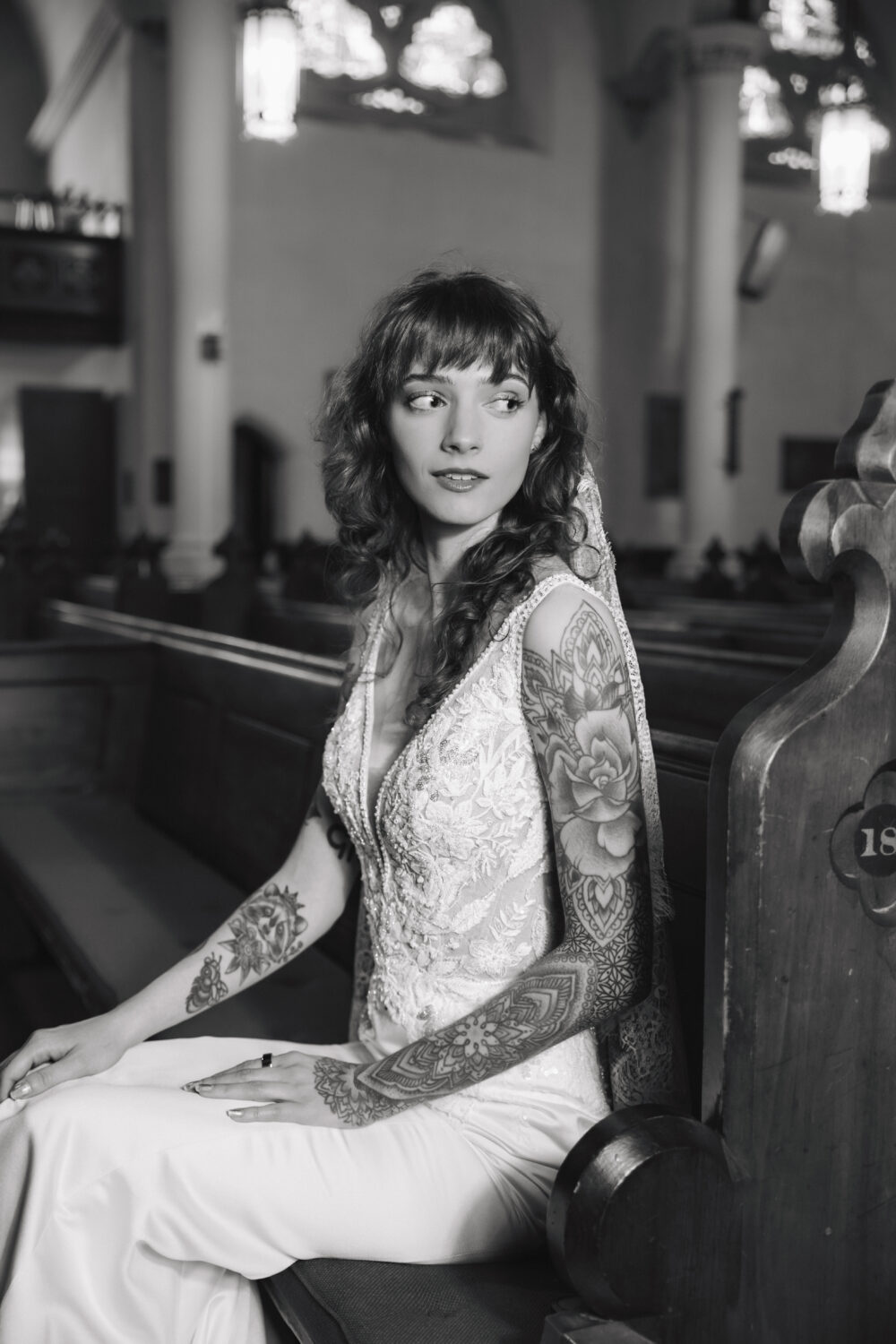 A bride with wavy hair and tattooed arms wears a lace gown and veil while sitting in a wooden church pew.