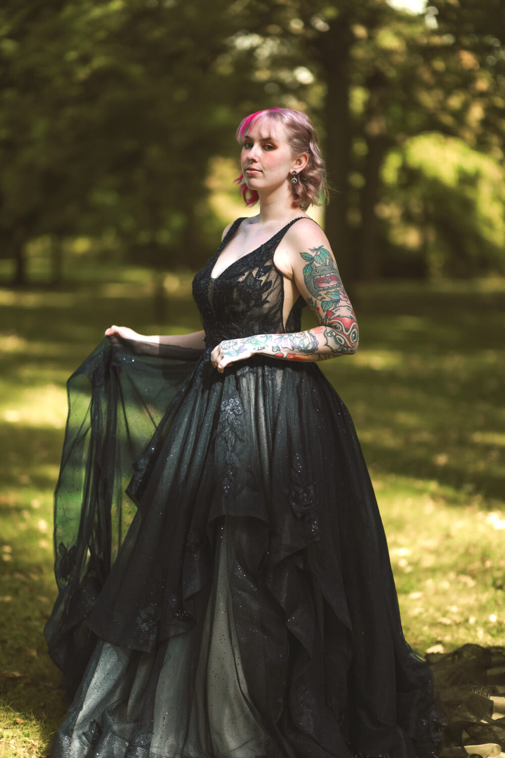 A woman with pink hair and tattooed arms poses outdoors in a black gown with a layered tulle skirt, holding the dress out to the side.