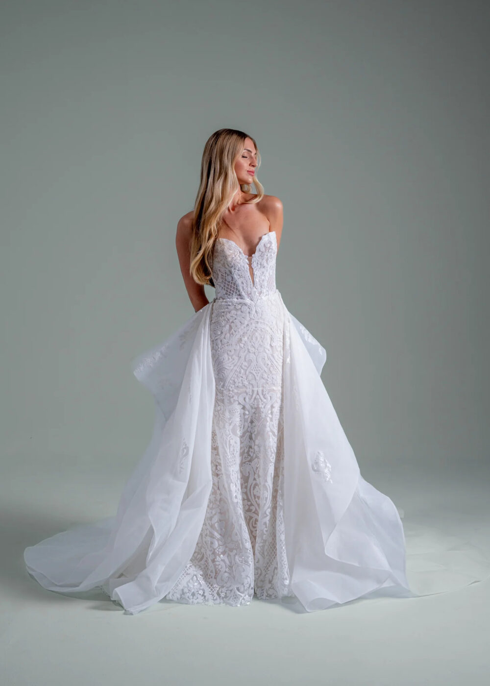 A woman wearing a white wedding dress has her eyes closed and both her hand on her back.