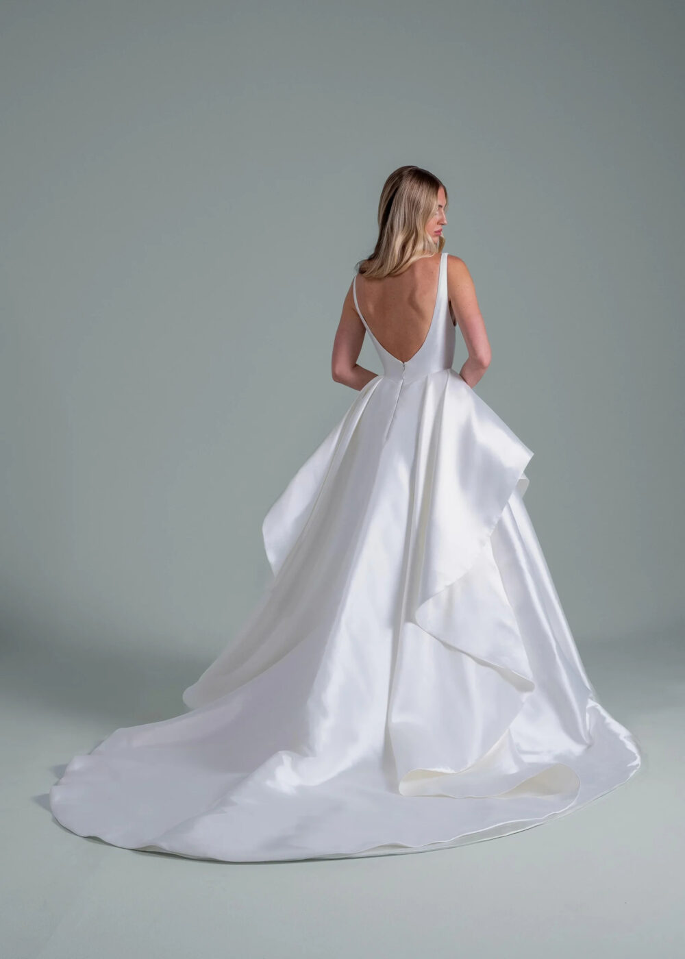 A woman in a sleeveless white satin gown with a deep V-back and cascading layered train stands facing away against a plain grey background.