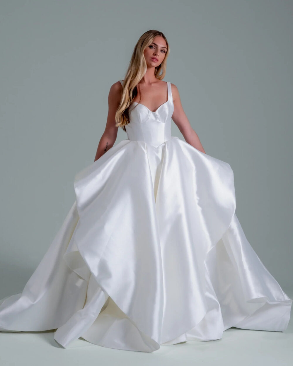 A woman wearing a white satin ball gown with wide shoulder straps, a fitted bodice, and a full layered skirt poses against a plain grey background.