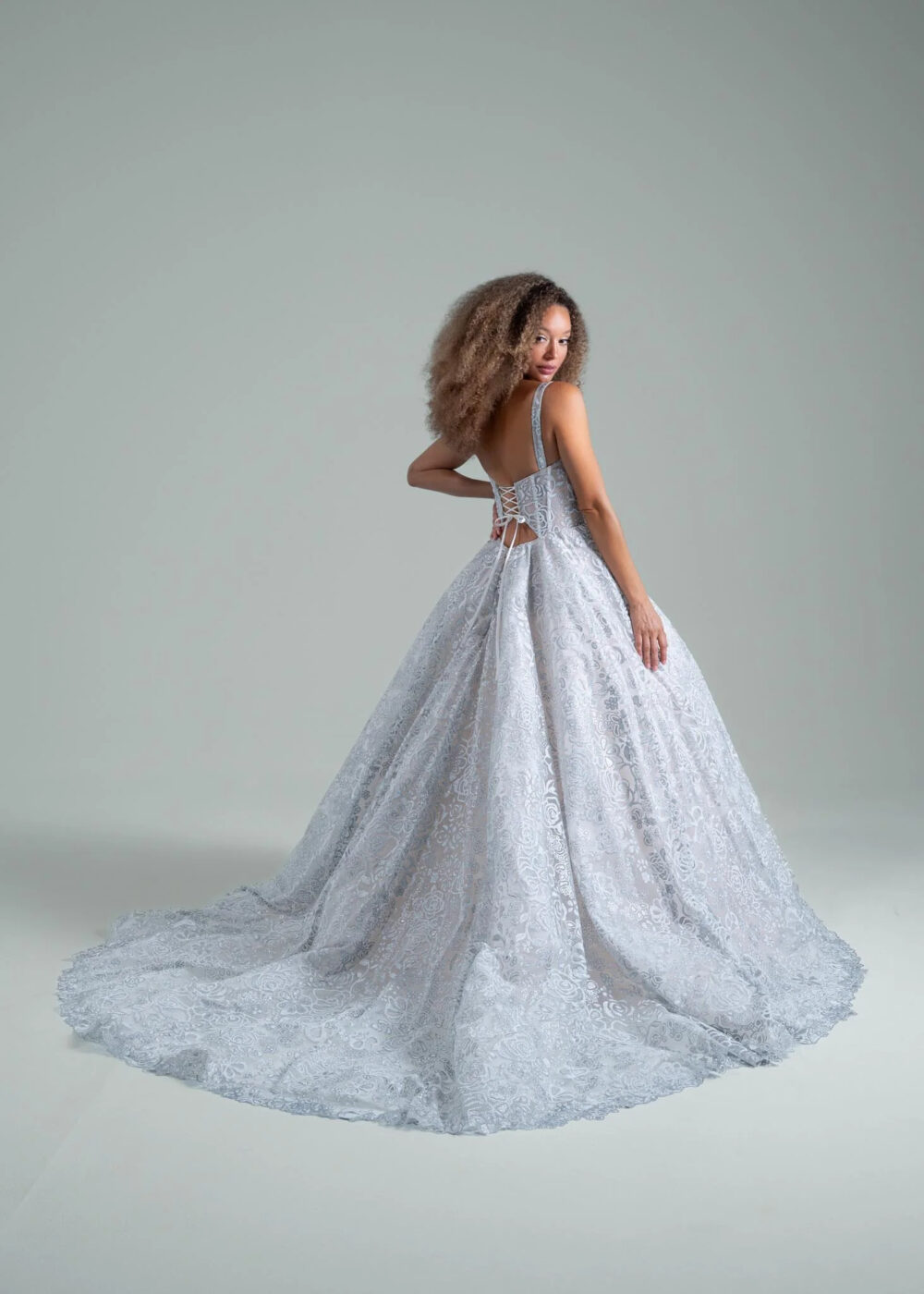 A woman wearing a white lace ball gown with a low back, lace-up corset detail, and a sweeping floral-patterned train poses while looking over her shoulder against a plain grey background.