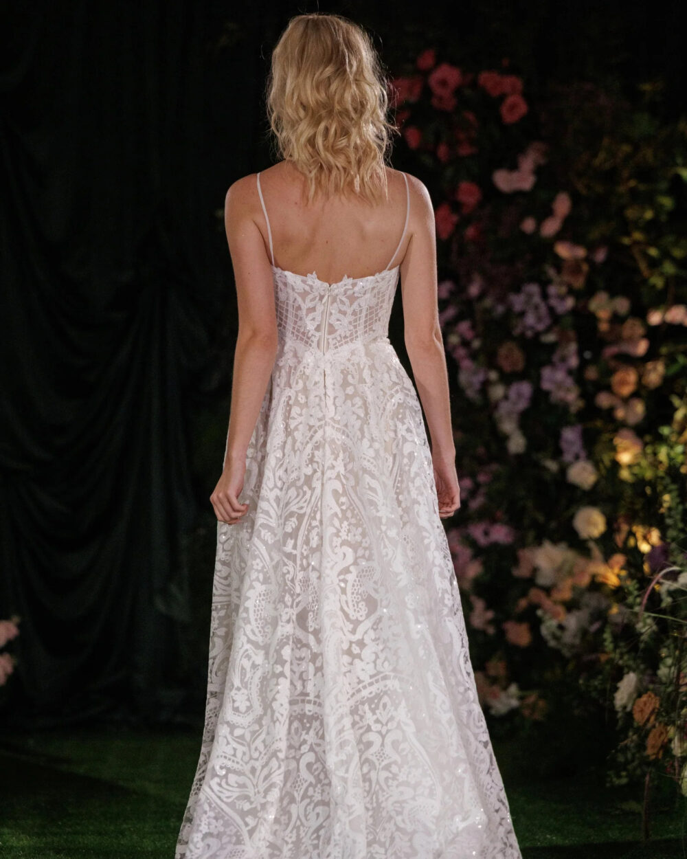 A woman wearing a lace A-line dress walks down a runway with a black backdrop designed with colorful flowers.