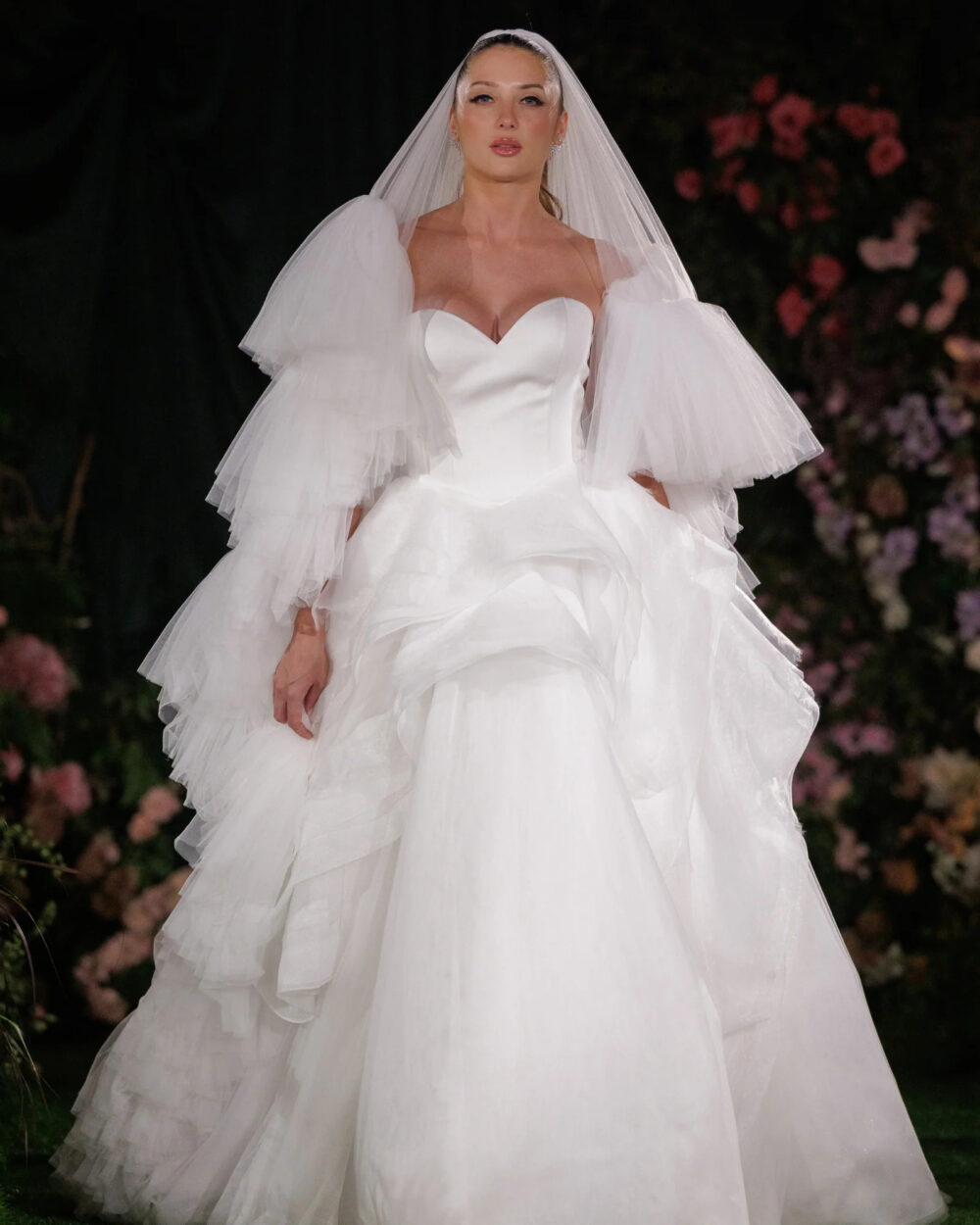 A bride in a strapless white satin gown with a sweetheart neckline, layered ruffled sleeves, and a long flowing veil walks down the runway against a backdrop of dark drapery and flowers.
