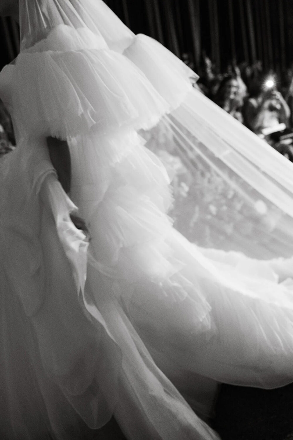 Black and white close-up of a bride’s layered tulle gown and veil, highlighting the voluminous ruffles as she walks down the runway