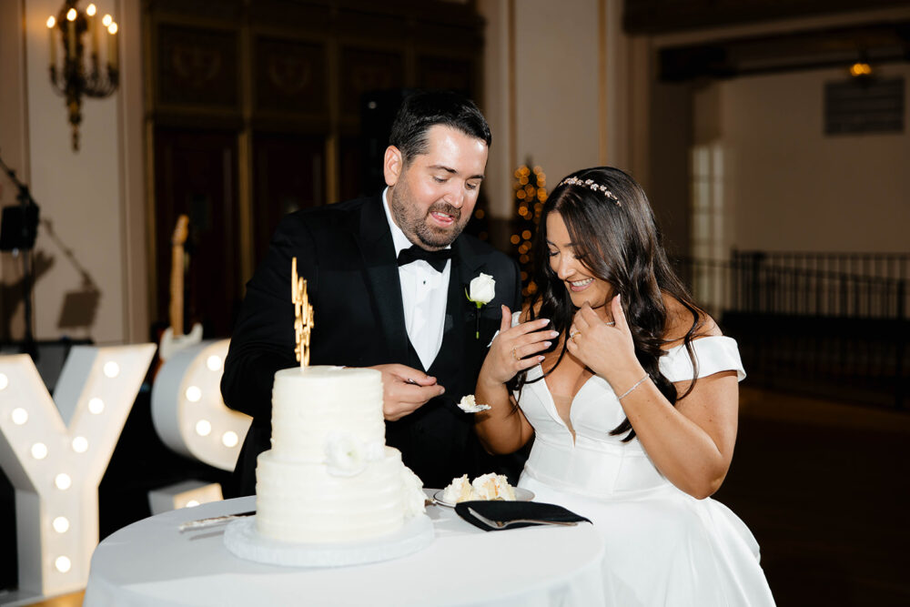 A bride and groom laugh while feeding each other wedding cake beside a two-tier white cake on a round table.